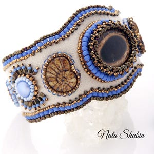 May include: A white felt bracelet with blue and brown beads, featuring a large brown eye-shaped stone and two smaller shell-like stones. The bracelet is decorated with a beaded border.