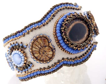 May include: A white felt bracelet with blue and brown beads, featuring a large brown eye-shaped stone and two smaller shell-like stones. The bracelet is decorated with a beaded border.