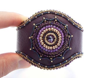 May include: A purple leather cuff bracelet with a beaded mandala design. The mandala features a central crystal surrounded by gold and purple beads.