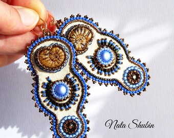 May include: A pair of white felt earrings with blue and brown bead embroidery. The earrings feature a circular design with a blue pearl in the center and a brown ammonite fossil.