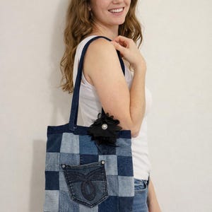 May include: A patchwork denim tote bag with a pocket and a black flower embellishment. The bag is made from various shades of blue denim, creating a checkered pattern. The tote has dark blue handles and a pocket from a pair of jeans.