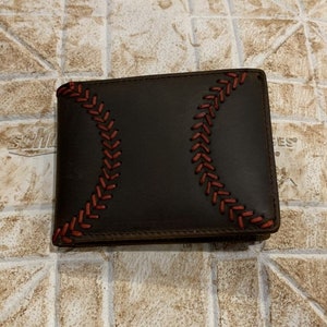 Pu&ograve; includere: Un portafoglio in pelle marrone con cuciture rosse a forma di palla da baseball. Le cuciture formano un motivo a palla da baseball.