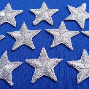 May include: Ten silver star-shaped iron-on patches on a blue background. The stars are approximately 2.5 cm in diameter.