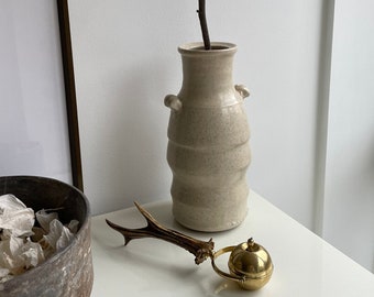 May include: A white ceramic vase with a textured surface and two handles holds a dried branch with twigs. A gold metal antler-shaped bottle stopper with a round, gold metal top sits on a white surface.