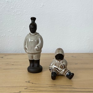 May include: Two ceramic figurines of people wearing hooded jackets. The figurines are brown and white and are standing on a wooden surface.