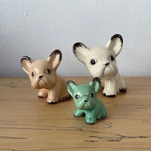 May include: Three ceramic figurines of French Bulldogs. The largest figurine is white with brown ears and a black nose. The middle figurine is light brown with brown ears and a black nose. The smallest figurine is green with brown ears and a black nose.