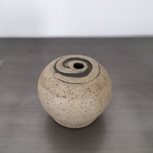 May include: A speckled, spherical ceramic vase with a spiral design around the top. The vase is a light beige color with a dark brown spiral and a hole in the center. The vase is sitting on a gray surface.