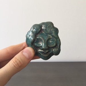 May include: A teal green ceramic sculpture of a face with a whimsical expression. The face has large eyes, a wide smile, and a textured surface.