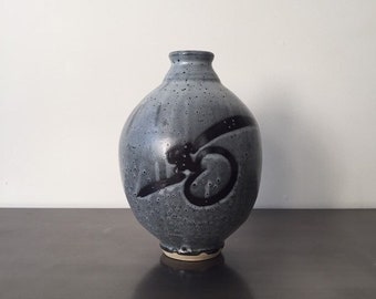 May include: A small, round, gray ceramic vase with a black abstract design. The vase has a speckled glaze and a slightly uneven base.