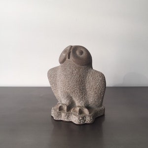 May include: A grey stone owl sculpture with detailed feathers and large eyes. The owl is perched on a base with a rough texture.