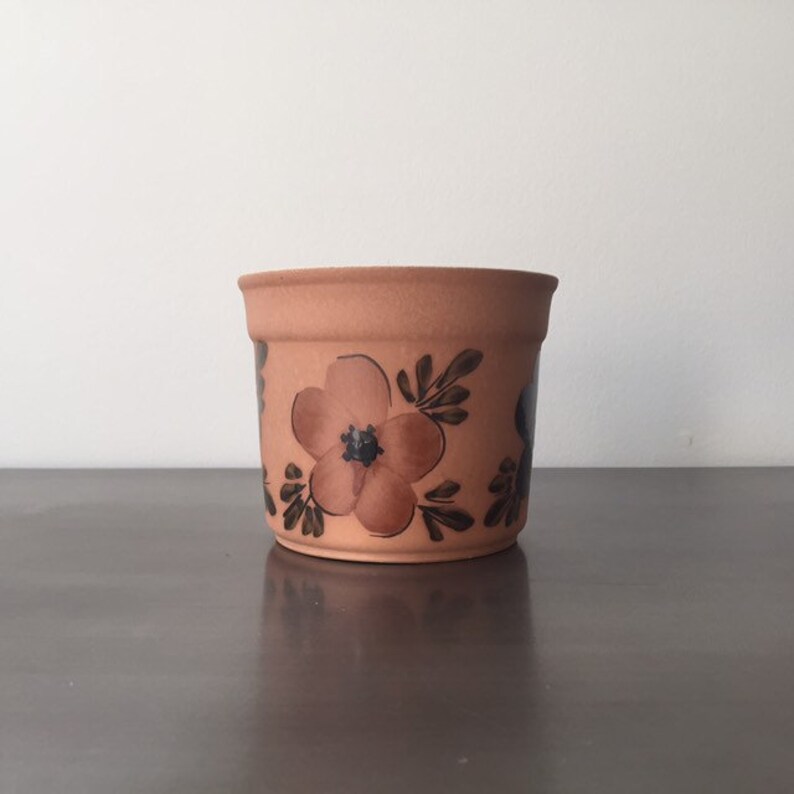 May include: A terracotta flower pot with a brown and black floral design. The pot is decorated with a large flower in the center and smaller flowers around the sides.
