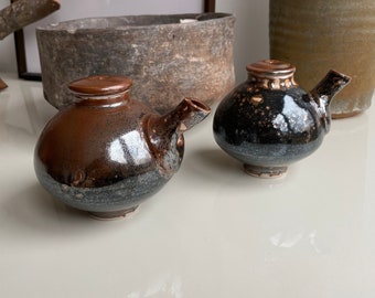 May include: Two ceramic teapots with brown and black glaze. The teapot on the left has a round body and a spout on the side. The teapot on the right has a round body and a spout on the top.