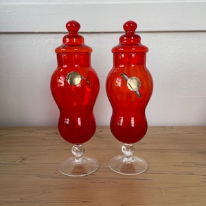 May include: Two orange glass jars with clear glass bases and lids. The jars have a round, bulbous shape and are decorated with a gold seal.