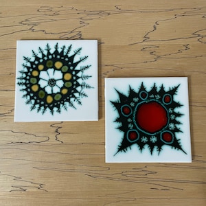 May include: Two white ceramic tiles with black and green abstract designs. The design on the left tile features a circular pattern with yellow and green accents. The design on the right tile features a red circle with black and green accents.