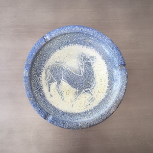 May include: A blue and white ceramic ashtray with a stylized horse design. The ashtray has a textured surface and a blue rim.