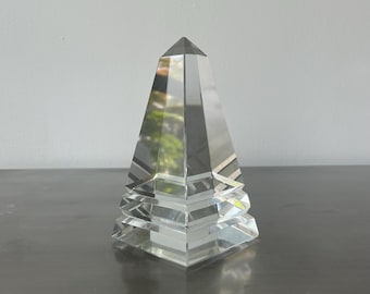May include: A clear, faceted crystal pyramid sculpture. The pyramid has a pointed top and a tiered base. The crystal reflects light, creating a shimmering effect. The sculpture is a decorative item.