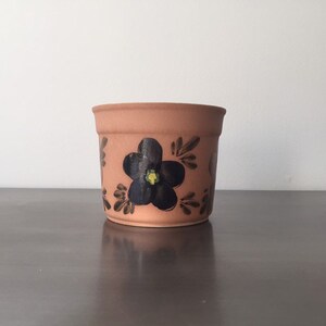May include: A terracotta flower pot with a black flower painted on the side. The flower has a yellow center.