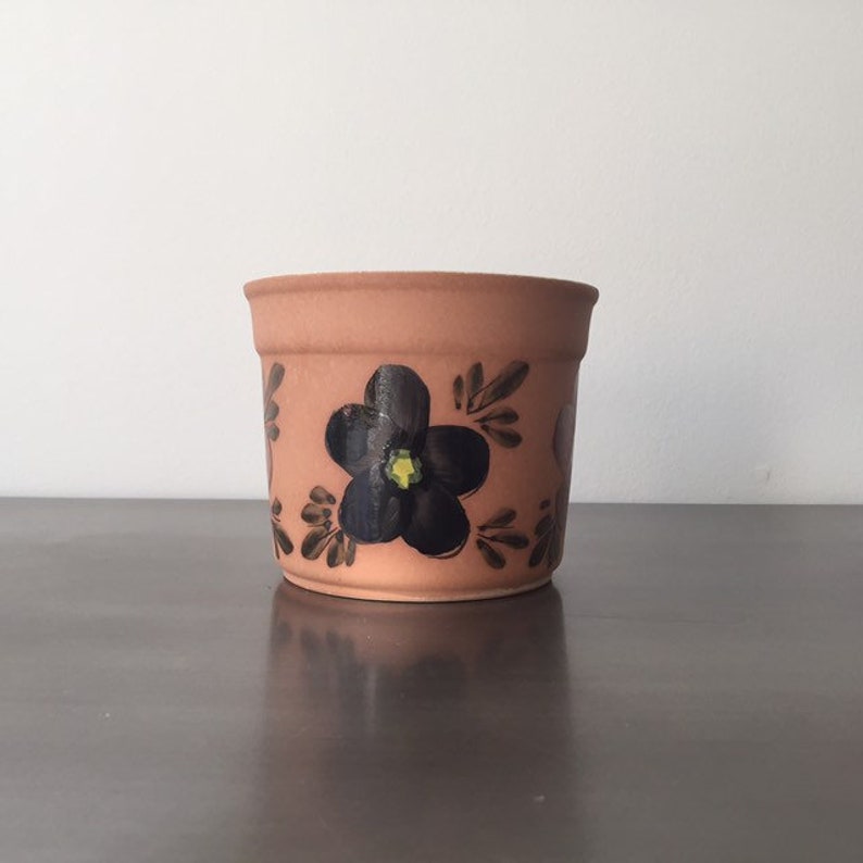 May include: A terracotta flower pot with a black flower painted on the side. The flower has a yellow center.
