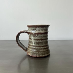 May include: A brown and white ceramic mug with a textured glaze. The mug has a wide, rounded handle.