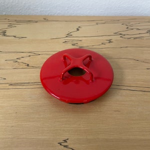 May include: Red metal lid with a circular shape and a cross-shaped handle. The lid has a small hole in the centre.