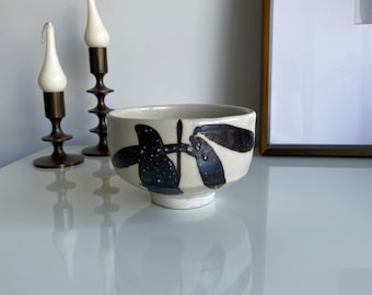 May include: A white ceramic bowl with black and brown abstract leaf designs. The bowl is sitting on a white surface.