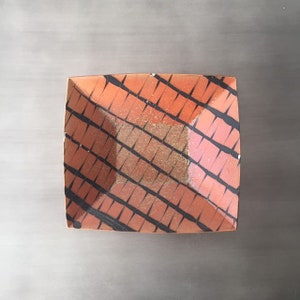 May include: A square ceramic dish with a black and orange geometric pattern. The dish has a slightly raised rim and a textured surface.