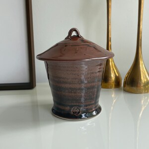 May include: A brown ceramic jar with a lid and a handle. The jar has a dark brown glaze with a lighter brown stripe pattern. The lid has a small knob on top.