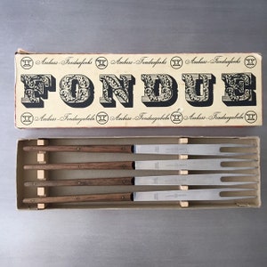 May include: A set of four fondue forks with wooden handles in a box. The box is white with black lettering that reads "Amboss-Fondueforks" and "FONDUES".