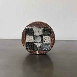 May include: A circular ceramic bottle stopper with a geometric design. It features a black and white checkerboard pattern, a silver-coloured circle in the centre, and a brown outer rim.