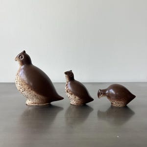 May include: Three ceramic bird ornaments in varying sizes. The birds are brown with speckled beige bellies and have a smooth, glossy finish. The ornaments are arranged in a row, showcasing their unique shapes and details.