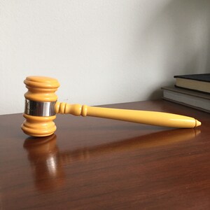 May include: A yellow wooden gavel with a silver band around the head. The gavel is lying on a wooden surface.