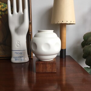 May include: White ceramic vase with a textured, moon-like surface. The vase is sitting on a dark wood base.