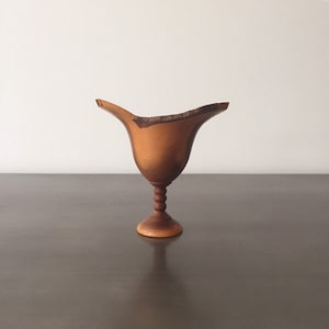 May include: A wooden vase with a unique, asymmetrical design. The vase is made of light brown wood with a darker brown base. It has a wide, open top and a narrow, rounded base.