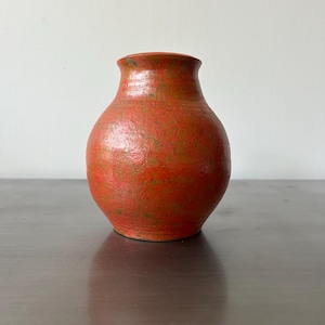May include: A small, round, orange ceramic vase with a textured surface. The vase has a narrow neck and a slightly flared rim. Greenish-brown speckles add detail to the orange colour. The vase sits on a dark surface.