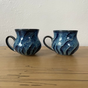 May include: Two ceramic mugs with a unique, textured design. The mugs feature a dark blue glaze with lighter blue and black accents, creating a wave-like pattern. Each mug has a rounded body and a curved handle. The mugs are displayed on a wooden surface.