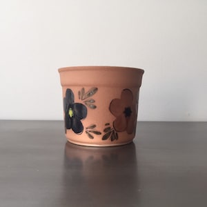 May include: A terracotta flower pot with hand-painted black and brown flowers. The pot is a light brown color and has a smooth, slightly textured surface.