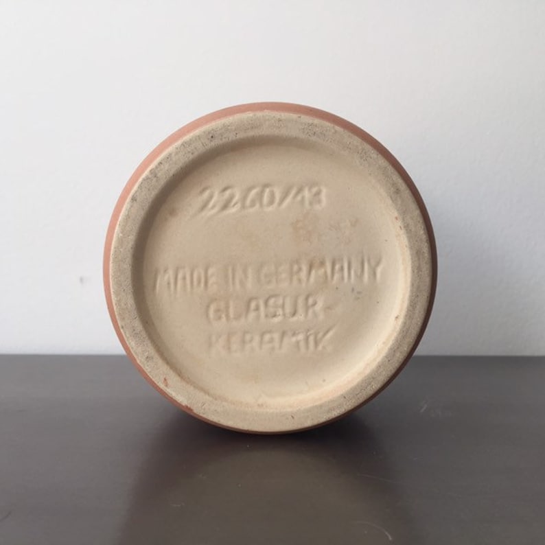 May include: The bottom of a terracotta-colored ceramic vase with a white glazed surface. The vase has the text "22.CD/13 Made in Germany Glasur-Keramik" printed on the bottom.