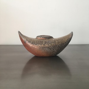 May include: A ceramic, crescent-shaped decorative bowl with a speckled, textured surface. The bowl features a gradient of colors, transitioning from a reddish-brown base to a speckled brown and tan top. A small, circular detail sits atop the center.