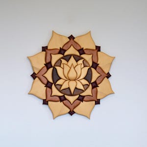 May include: A wooden lotus flower wall art piece with a light brown and dark brown color scheme. The art is mounted on a white wall. Other items on a wooden surface include a framed picture, a gray towel, and a decorative vase.