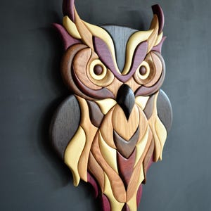 Peut inclure: Une œuvre d'art murale en forme de hibou en bois. Le hibou est composé de différentes teintes de bois, notamment marron foncé, marron clair, jaune et bordeaux. L'œuvre est montée sur un fond gris foncé.