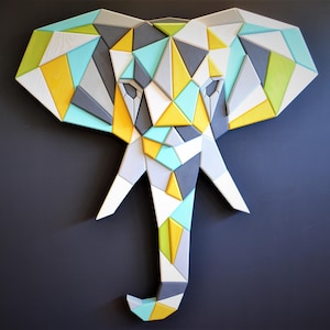 Op de afbeelding: Geometrische houten olifantenkop wandkunst in tinten geel, grijs, wit en blauwgroen. De olifant is opgebouwd uit verschillende driehoekige en rechthoekige stukken, wat een modern, abstract ontwerp creëert. Het kunstwerk is op een donkergrijze muur gemonteerd.