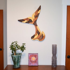May include: A wooden whale tail wall art piece with a mosaic design in shades of brown, tan, and black. The art is mounted on a white wall. The piece is a decorative item for a nautical or coastal themed home.