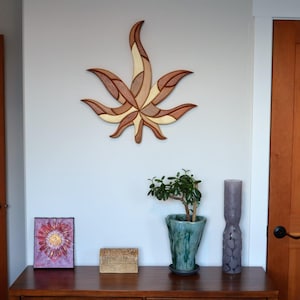 May include: A wooden wall art piece in the shape of a cannabis leaf, with various shades of brown and tan. Below, a wooden cabinet displays a small potted plant, a purple candle, and a framed artwork.