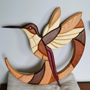 May include: Wooden hummingbird wall art. The bird is crafted from various wood tones, including light beige, medium brown, and dark brown. The hummingbird is depicted in flight, with its wings spread. The design includes a crescent-shaped base.