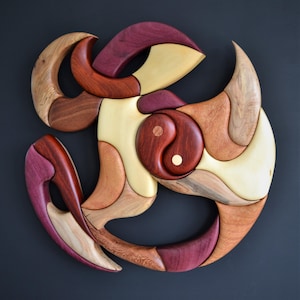 May include: A wooden wall art piece featuring an abstract design with various wood tones. The artwork incorporates a yin-yang symbol at the center, with curved and flowing shapes in shades of red, brown, and cream. The piece is mounted on a dark gray background.