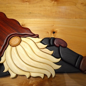 Op de afbeelding: Houten kabouterfiguur in een liggende houding. De kabouter heeft een donkerrode hoed, een lichte houten baard en een zwarte broek. De laarzen en de neus zijn bruin. Het figuur is gemaakt van diverse houten stukken.