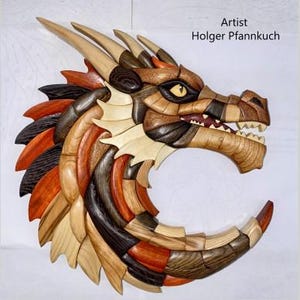 May include: A wooden dragon head sculpture with a detailed mosaic design. The dragon features a variety of wood tones, including light beige, brown, and reddish-orange. The artist's name, Holger Pfannkuch, is displayed in the upper right corner.