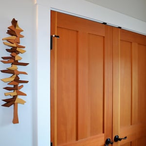 May include: A wooden wall art piece in the shape of a tree, composed of various geometric shapes in shades of brown and tan. The art is mounted on a white wall, next to a set of wooden double doors.