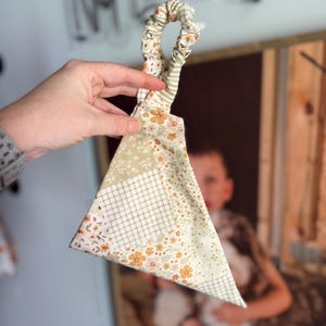 May include: A handmade fabric headband with a triangular design. The headband features a mix of floral and plaid patterns in beige, cream, and orange. The elastic loop is striped in beige and green.