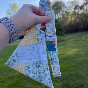 Puede incluir: Un conjunto de collar y pañuelo para perro con estampado floral. El collar presenta una hebilla plateada y un diseño floral azul claro y amarillo. El pañuelo tiene un diseño de patchwork con cuadros vichy amarillos y estampados florales azules. El conjunto se sostiene sobre un fondo verde.